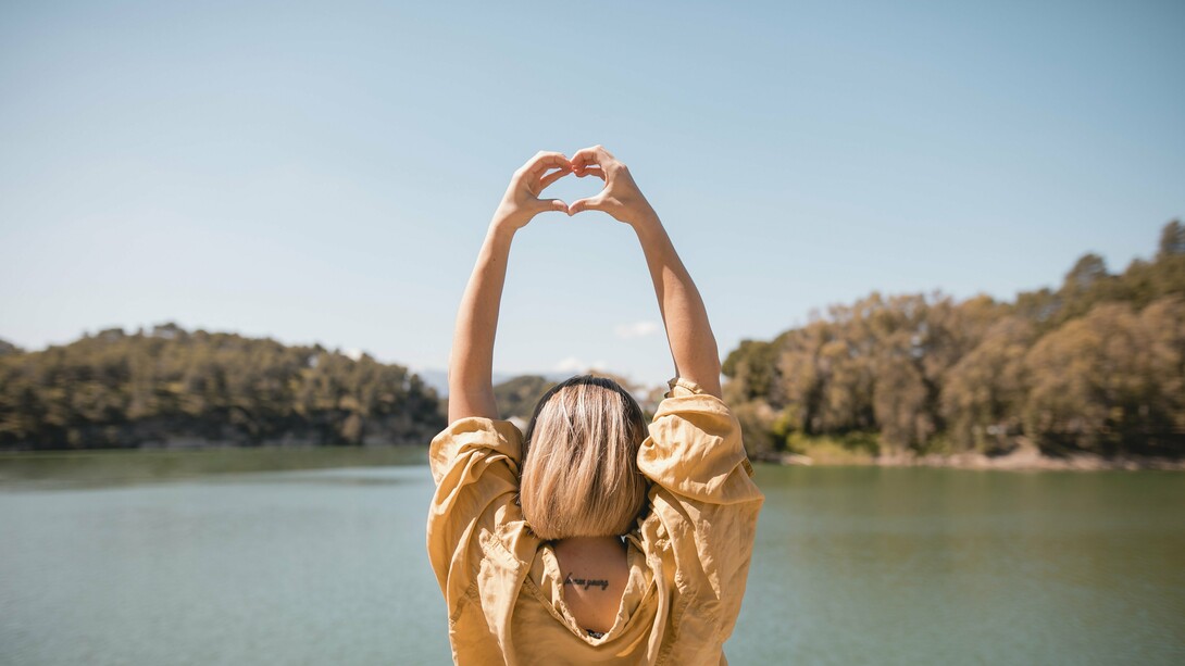 A woman by a serene lake, her arms extended above her head, forming a heart shape with her hands against a clear blue sky, an image that embodies the balance between nature, self-care, and resilience