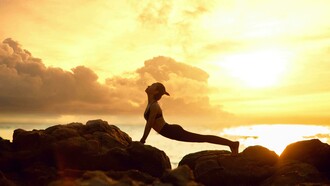 The silhouette of a woman practicing yoga on a rocky beach at sunrise evokes a sense of peace, balance, and deep relaxation
