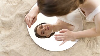 Una donna si china verso uno specchio rotondo sulla sabbia, riflettendo il suo viso