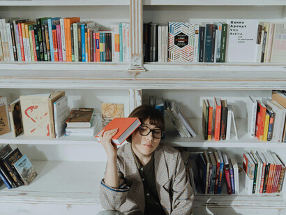 A young woman sitting besides a bookshelf, holding a book to her head