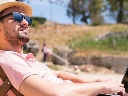 Flexible summer work setup: Man using a laptop from a beach chair with a drink, balancing productivity and relaxation