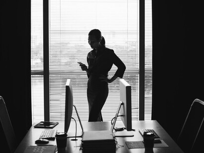 A dark silhouette of a woman standing by a window in an office, using her smartphone, representing modern business communication