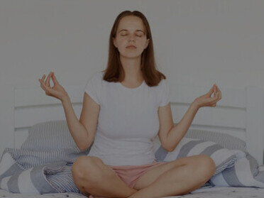 Indoor image of a dark-haired woman in a white t-shirt and shorts, seated in a lotus position on a bed in a bright, airy bedroom, smiling and meditating with palms pressed together in the morning