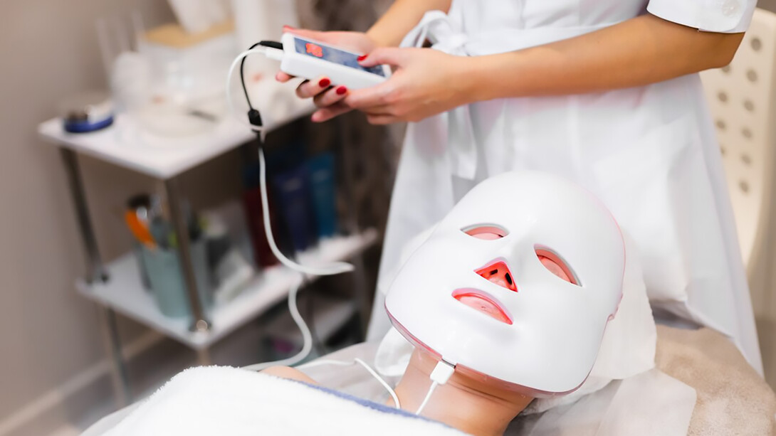 A client relaxes on a cosmetology table in a salon, wearing a white facial mask while enjoying the rejuvenating effects of red light therapy