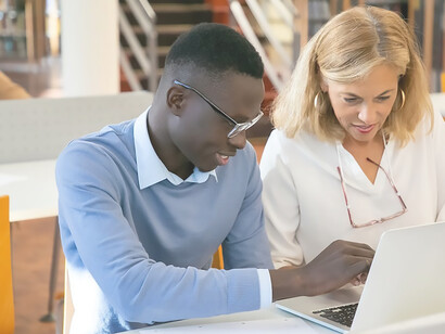 Young student using laptop while preparing for exam with his female professor in library