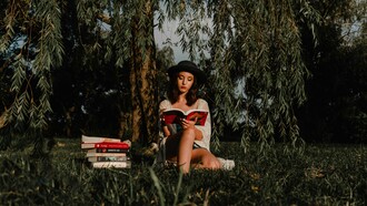 Jovem lê um livro sob a sombra de uma árvore, cercada por outros livros. A imagem remete ao início de uma busca interior que une contemplação, conhecimento e silêncio