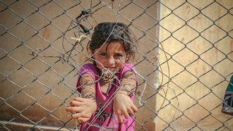 A young girl grips a chain-link fence in Idlib, Idlib Governorate, Syria