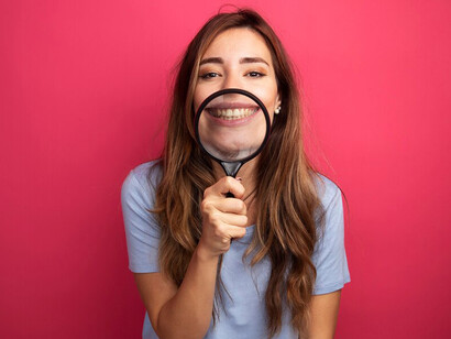 Young woman in a blue t-shirt, smiling and holding a magnifying glass to her mouth, against a pink background, highlighting oral health and hygiene
