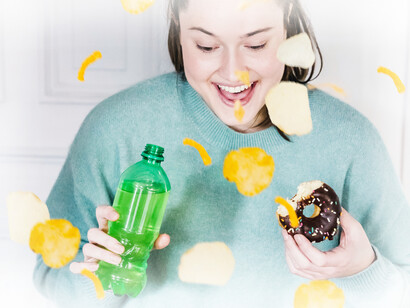 A woman smiles as chips and snacks fly in the air, drinking a sugar-filled soda and donut she is eating