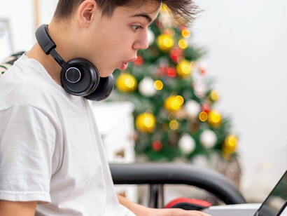 A teenage boy sits at home, immersed in a digital world as he uses a smartphone with headphones and balances a laptop on his knees, highlighting the influence of technology and the challenges of digital distractions for children