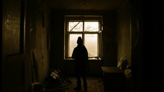 A solitary figure stands silhouetted by a window in an abandoned house, surrounded by fading light and quiet decay