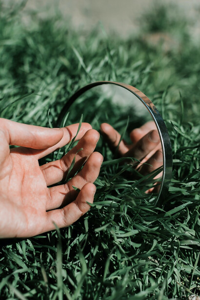 A person holds a black round frame, capturing the reflection of the surrounding grass and the open sky in its glass