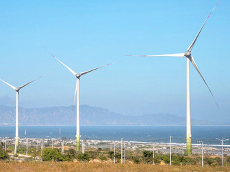 Offshore wind turbines stand tall against the vast ocean and rugged ridge under a clear blue sky, showcasing the UK's commitment to renewable energy amidst stunning natural landscapes