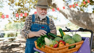 An elderly man wearing denim overalls and a hat, examining fresh vegetables