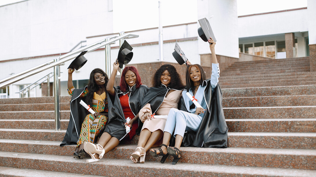 A picturesque scene unfolds as a group of young African American female students dons black graduation gowns against the backdrop of a campus setting