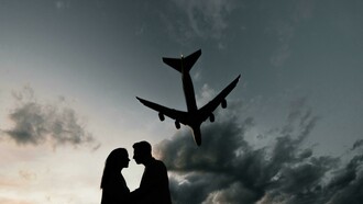 Silhouettes of a couple sharing a farewell embrace under a plane flying across the sky at the airport