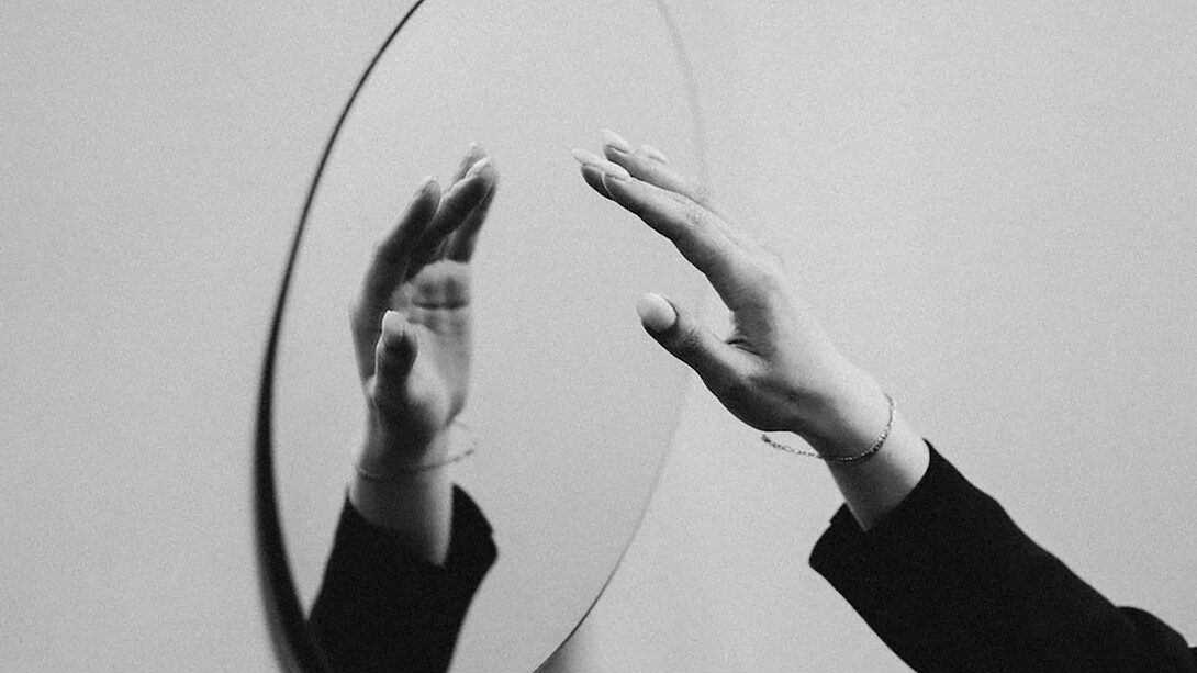 A person delicately reaches out to touch a mirror, and in this interaction, a reflection of her hand appears over the mirror's surface, creating an intriguing visual connection between reality and its mirrored counterpart