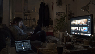 A young man sits alone in a dimly lit room, surrounded by screens, absorbed in a digital world