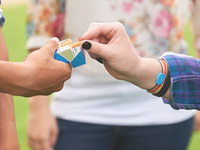 In a close-up shot, a teenage boy offers a cigarette to a friend of his