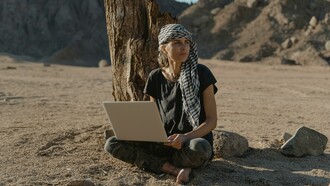 A woman in a black shirt sits on the ground in the desert, working remotely on her laptop