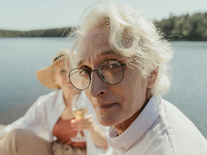 An eldery man with glasses looking at the camera, sitting on a pier with a woman, enjoying a sunny day