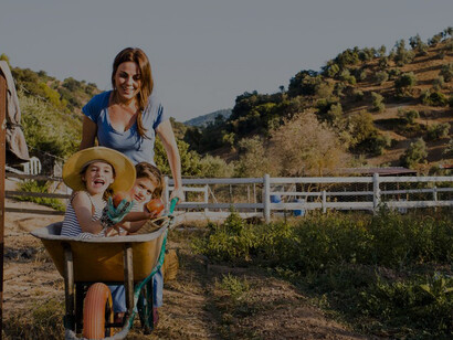 Happy family on rural European holiday, with mother joyfully pushing two daughters in wheelbarrow through scenic countryside