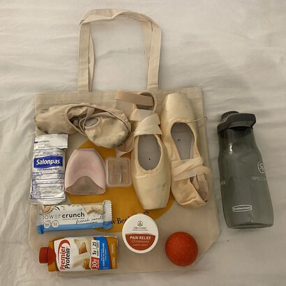 A ballet dancer's bag, filled with shoes, water, and a protein drink among other items
