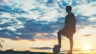 A young footballer silhouetted against the evening sky with a ball at their feet, representing dreams that reach far beyond the horizon