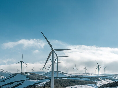Against the backdrop of a snow-covered field, windmills rise gracefully, harnessing the power of the daytime sun to generate renewable energy in a picturesque winter setting