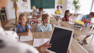 In un'aula scolastica, un'insegnante in primo piano tiene in mano un tablet, i suoi studenti sono seduti ai banchi