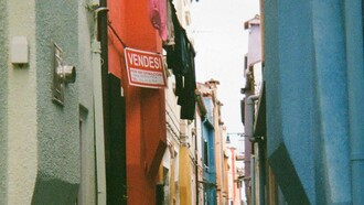 Un vicolo stretto a Burano, Italia, fiancheggiato da edifici dai colori vivaci, con un cartello rosso con la scritta "Vendesi" su uno degli edifici