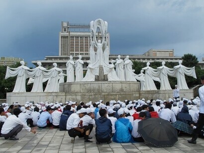 Raduno di fronte al Monumento della Fraternità: Persone sedute davanti a un imponente gruppo scultoreo che rappresenta l'unità e la collaborazione, un simbolo di coesione e speranza nel cuore di Pyongyang, Corea del Nord