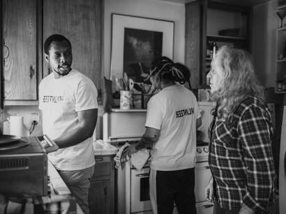 Two volunteers keep an elderly man company and help clean his kitchen