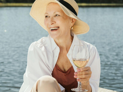 An eldery woman wearing a hat smiles and holds a glass of wine on a pier, taking on the sunlight